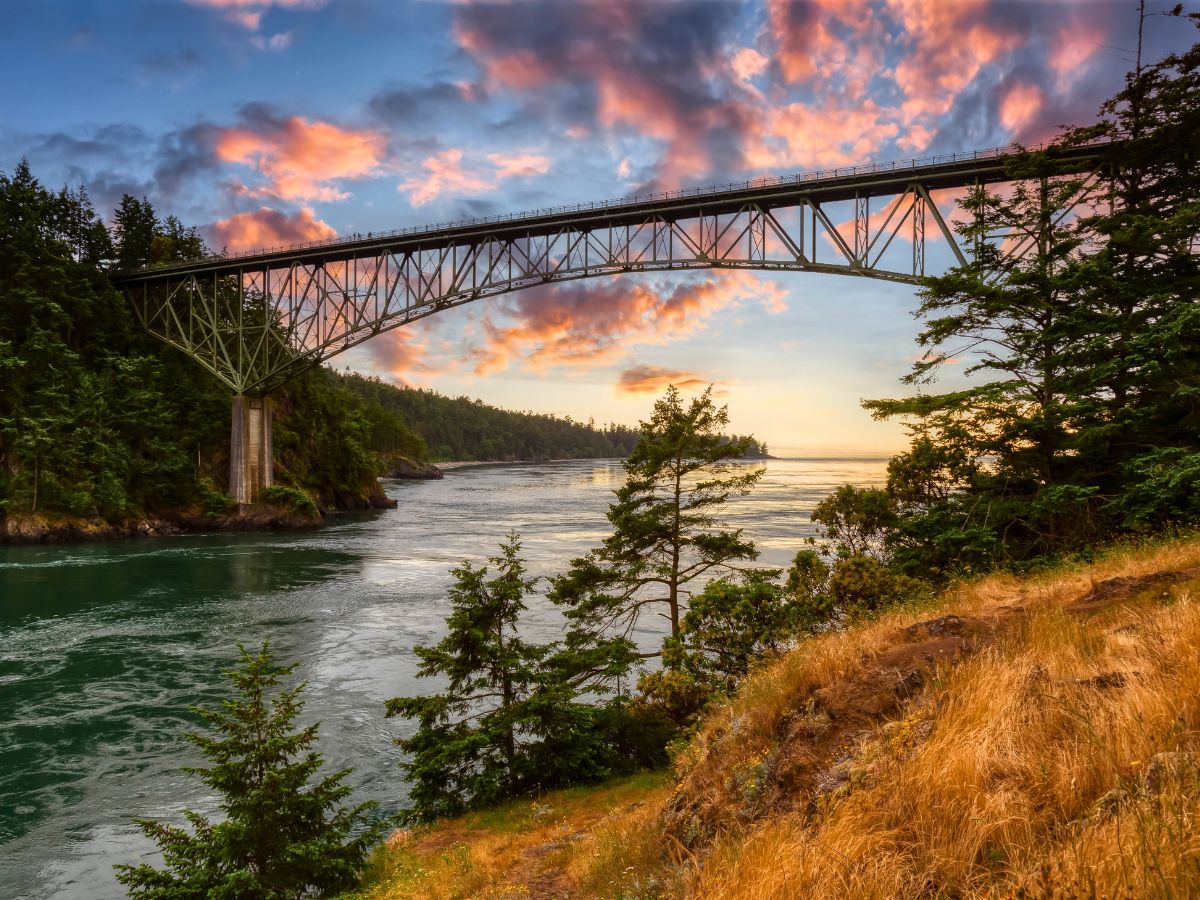A steel bridge spans a body of water, surrounded by trees and grassy hills, with a colorful sunset sky in the background.