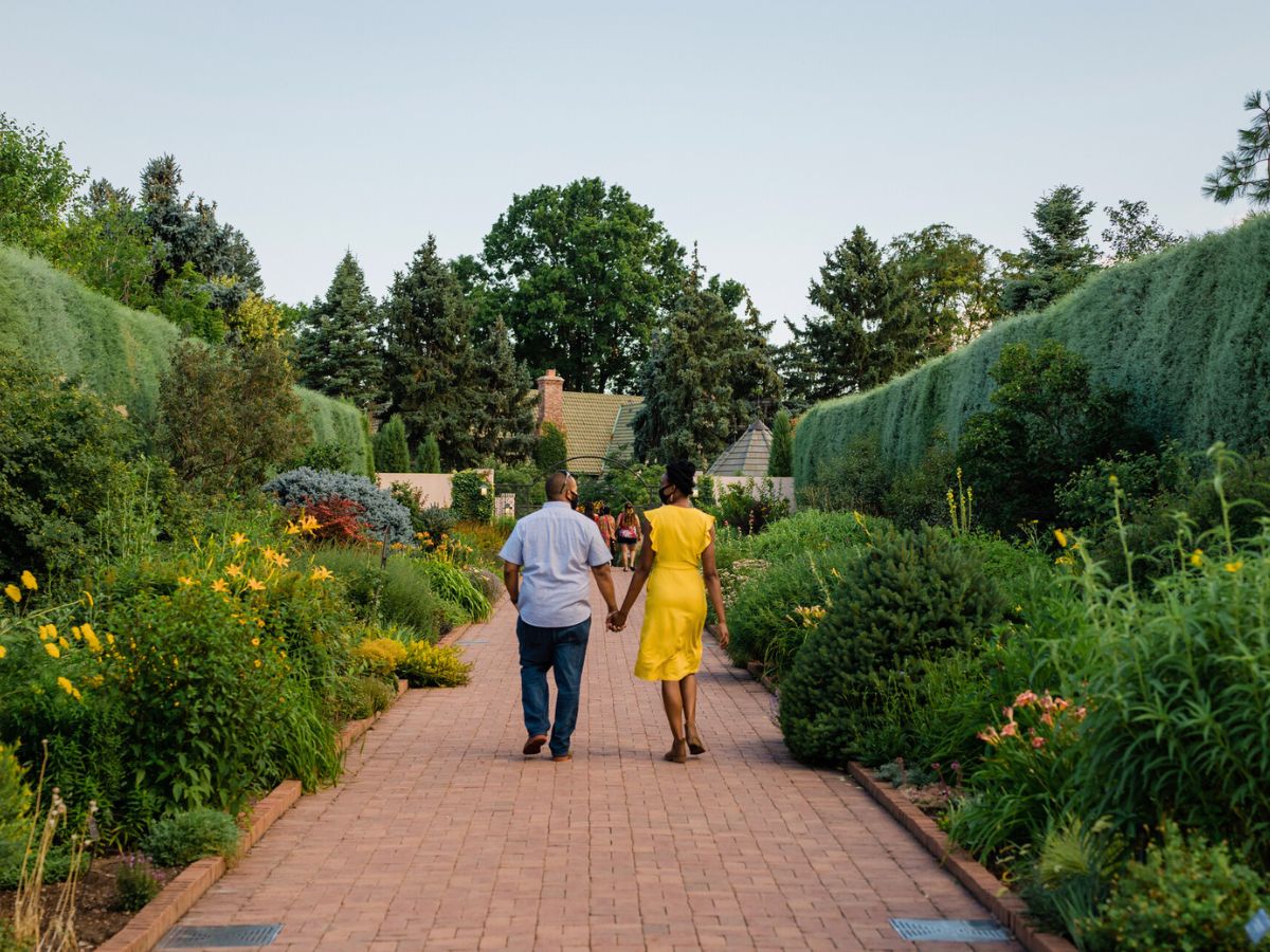A couple holds hands while walking down a brick path in a lush garden, surrounded by various green plants and colorful flowers.