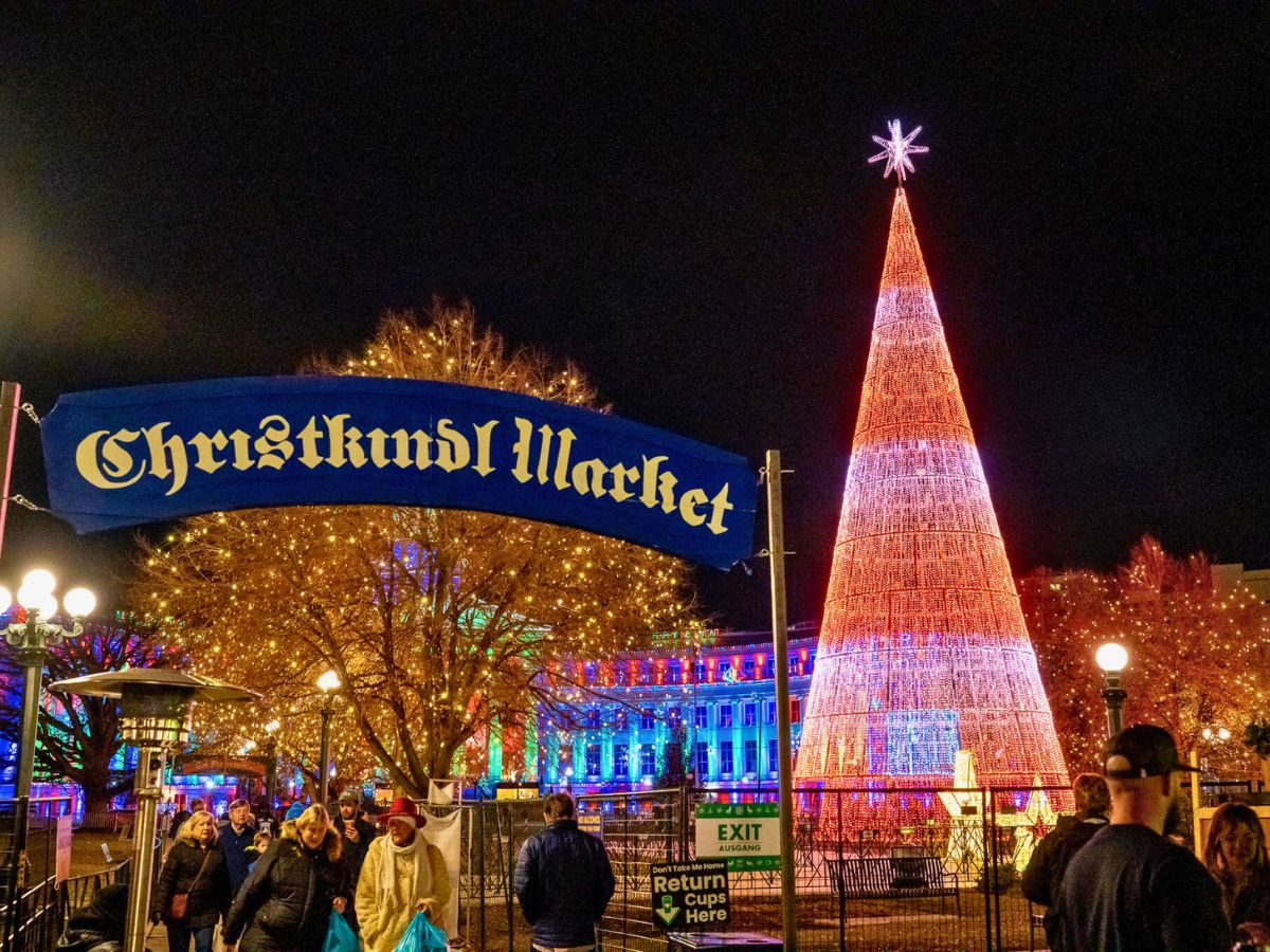 Festive Christmas market with bright lights and a large illuminated tree. Crowds explore the market at night.