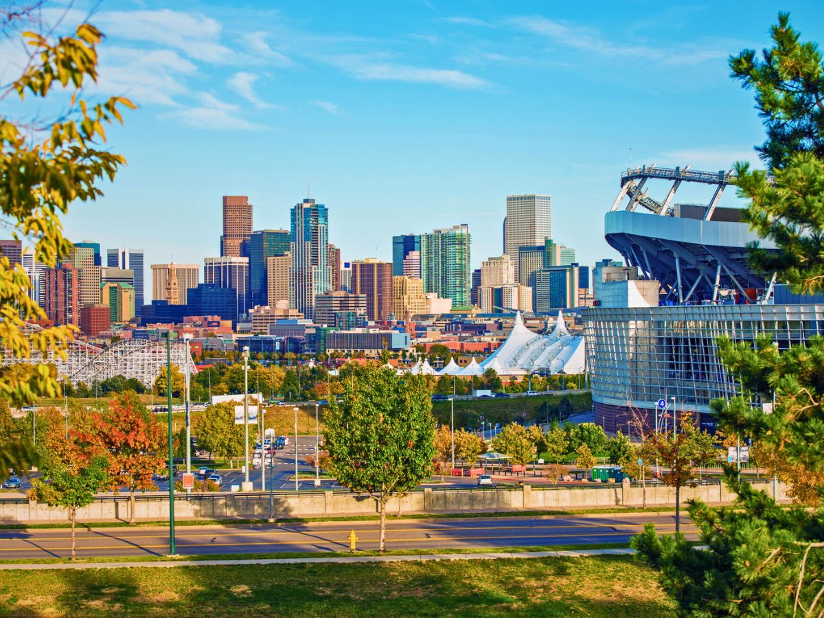 City skyline with modern buildings in the background, a large stadium on the right, and trees with colorful foliage in the foreground under a blue sky.