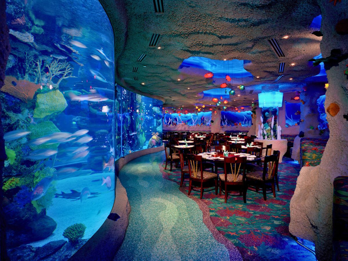 A dimly lit underwater-themed restaurant with large aquarium walls, colorful fish, and set dining tables.