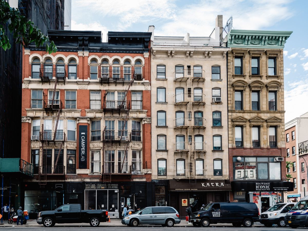 A street view shows five adjacent multi-story buildings with storefronts and fire escapes, and several parked cars and pedestrians on the sidewalk in an urban area.