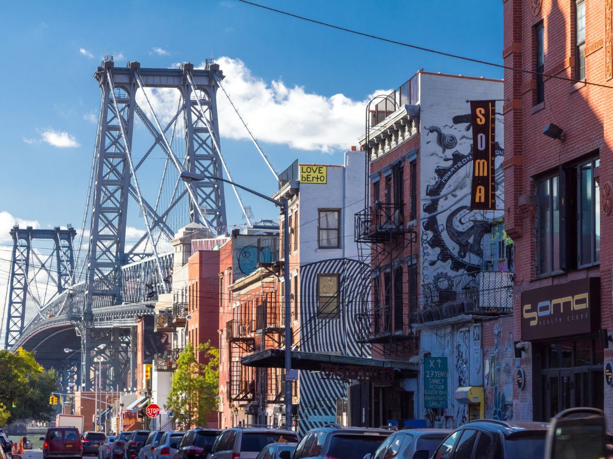 Street scene with buildings featuring murals and signs, with a large suspension bridge in the background under a blue sky. Cars are parked along the sides of the road.