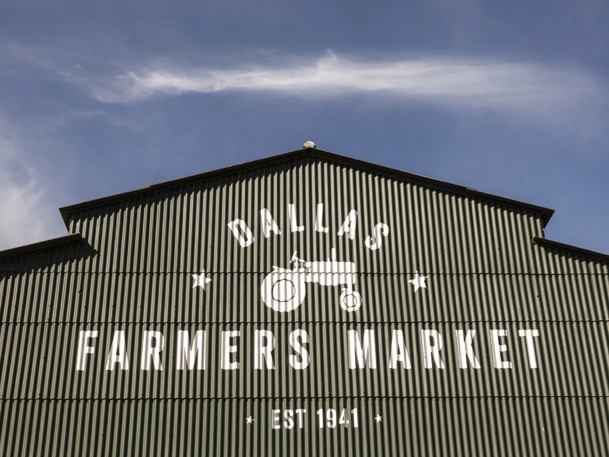 Green corrugated metal building with "Dallas Farmers Market Est. 1941" painted in white, featuring a tractor graphic, under a clear sky.