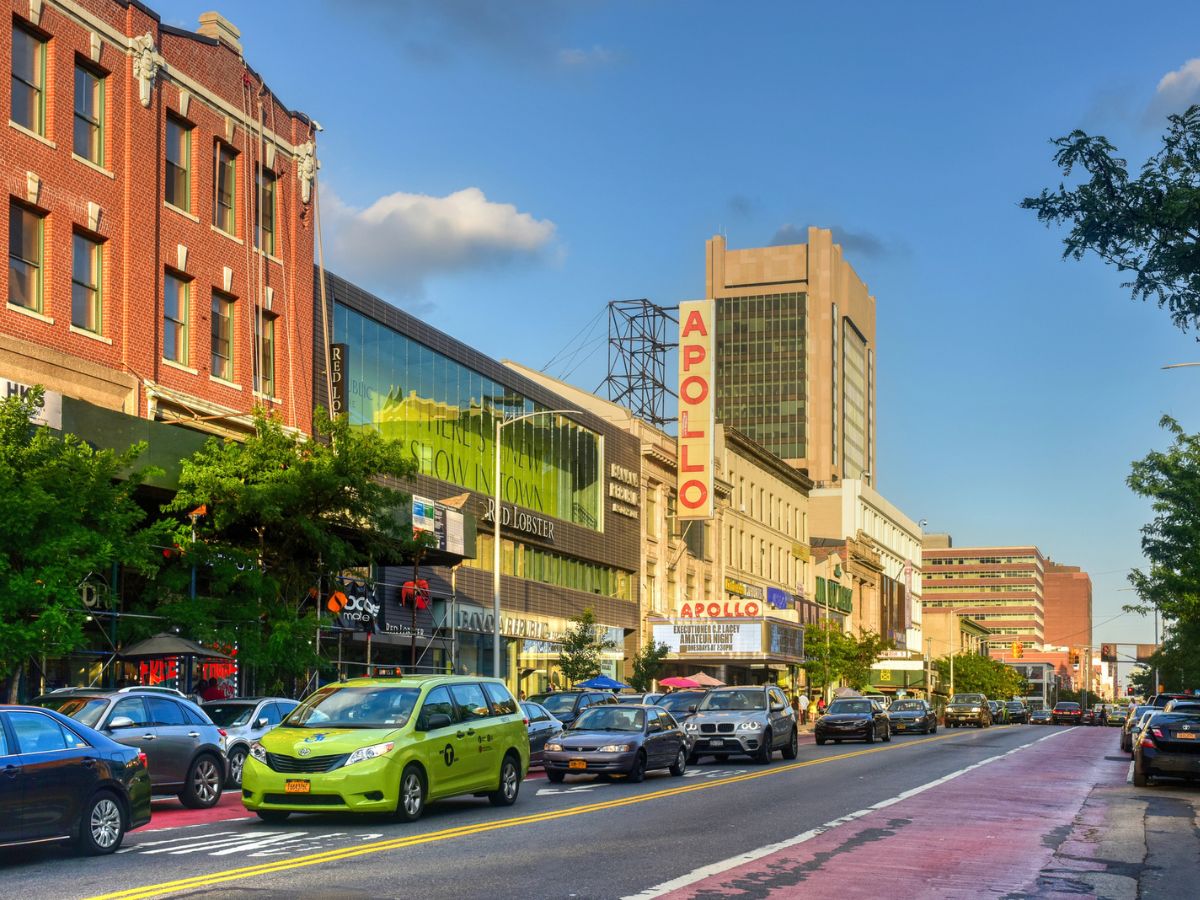 A busy city street with several cars and taxis. The Apollo Theater sign is prominently visible on one of the buildings. The street is lined with storefronts and trees under a clear sky.