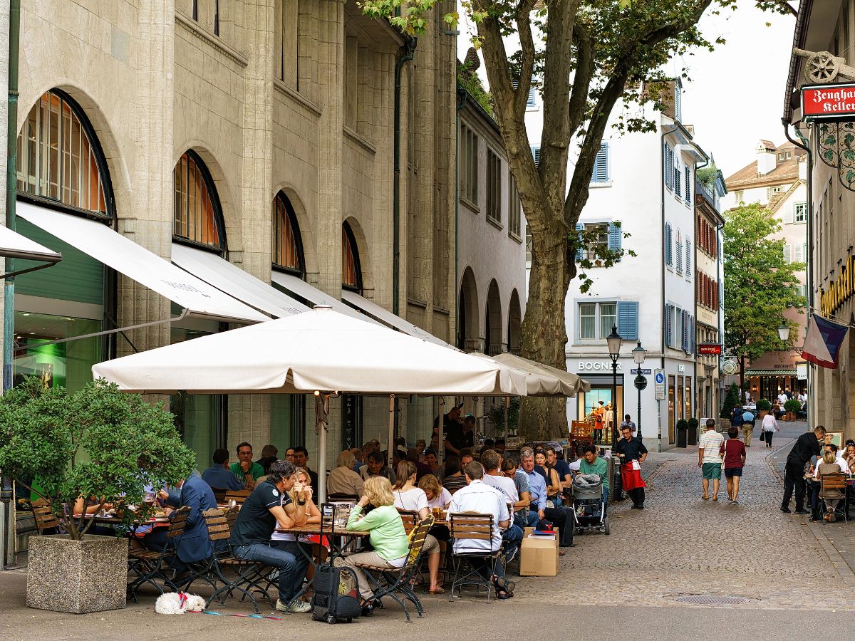 People sit at outdoor tables under umbrellas on a cobblestone street lined with shops and trees.