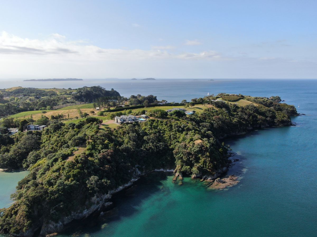 Aerial view of a coastal landscape featuring lush greenery, scattered buildings, and farmland on a cliff overlooking clear ocean waters with distant landmasses under a partially cloudy sky.