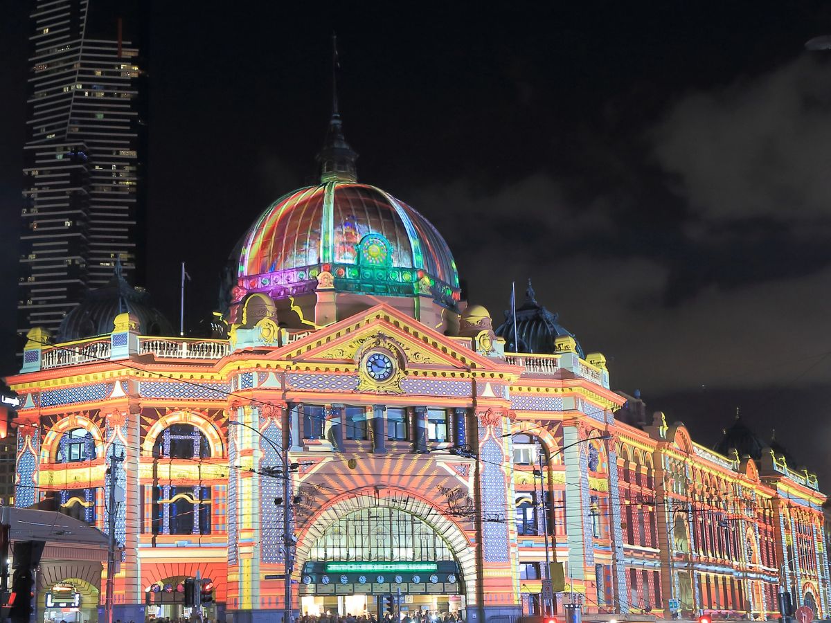 A brightly illuminated, historic train station with a colorful facade stands out against a nighttime city skyline. The building has a prominent dome and clocktower.