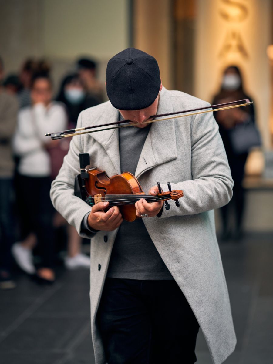 A person wearing a gray coat and black cap plays a violin with a bow held to their mouth in a public place, while onlookers in the background watch.