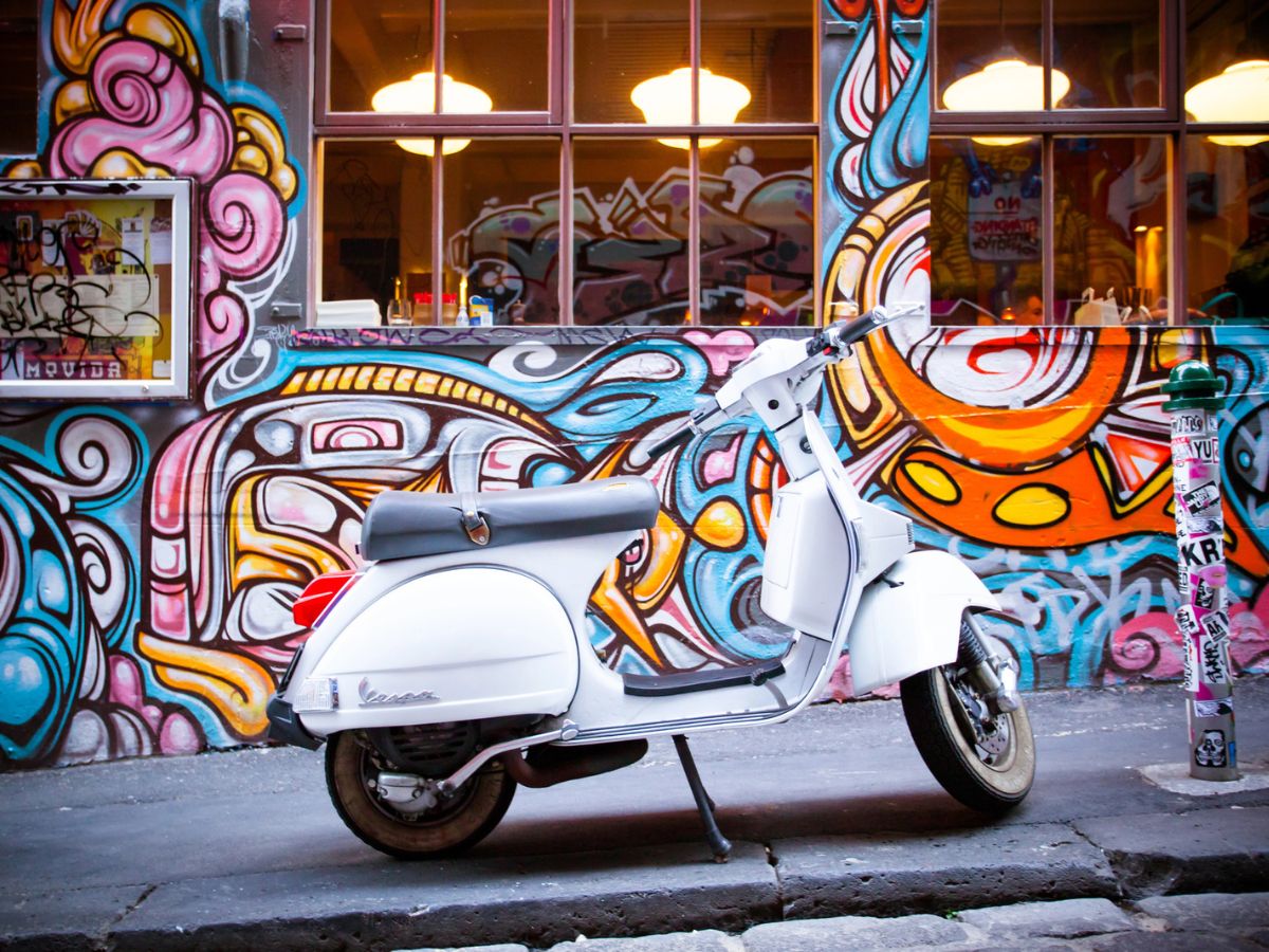 A white Vespa scooter is parked in front of a graffiti-covered wall with vibrant and colorful designs.