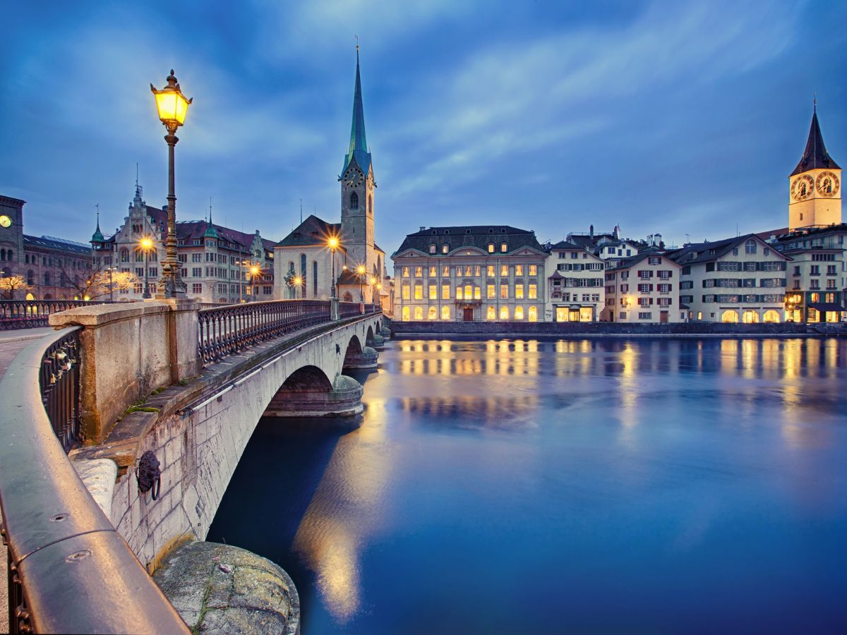 A serene cityscape of Zurich at dusk, featuring a bridge over the river, illuminated street lamps, and historic buildings against a cloudy sky.
