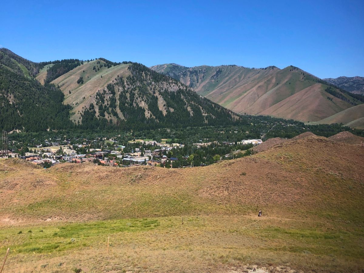 A small town surrounded by mountains under a clear blue sky, with grassy hills in the foreground.