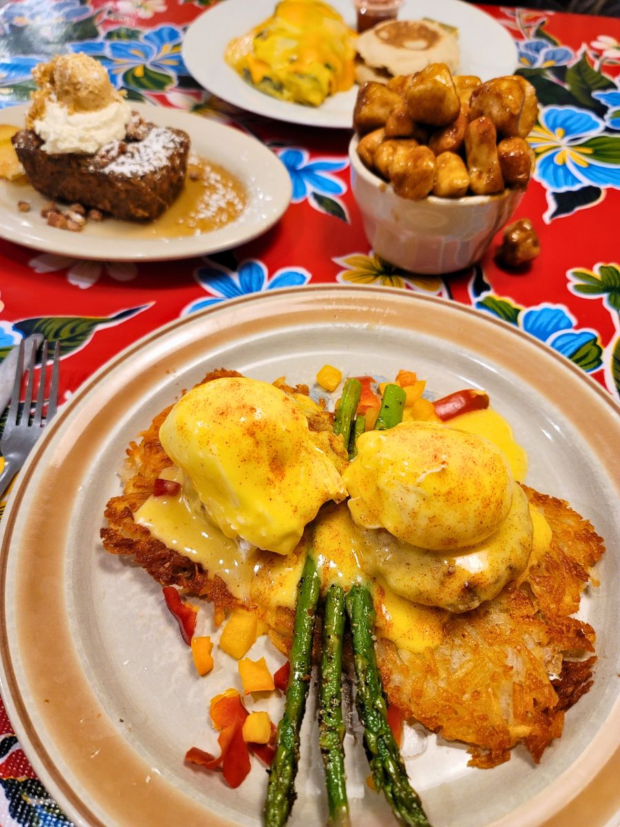 A plate of eggs Benedict on hash browns with asparagus, a dish of roasted potatoes, a dessert with whipped cream, and a plate with an omelette and biscuit on a colorful tablecloth.