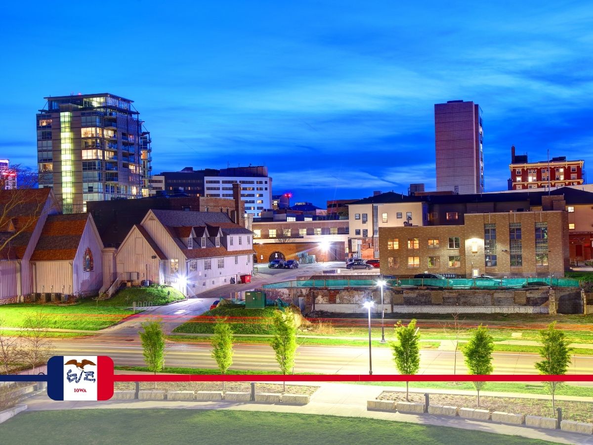Urban landscape at dusk showcasing various buildings, including a church and modern structures. The scene is under a clear blue sky, with the foreground featuring trees and a grassy area.