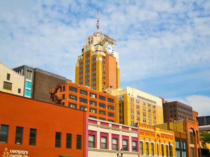 A cityscape featuring multiple colorful buildings, with a prominent clock tower in the background under a partly cloudy sky.