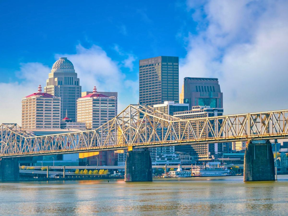 A city skyline with tall buildings is seen behind a large bridge spanning over a body of water under a blue sky.