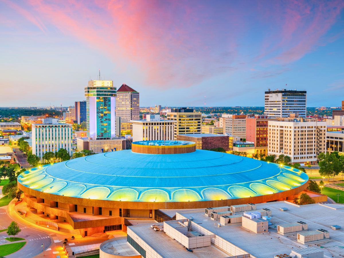 An aerial view of an urban skyline at sunset, featuring a large circular building with a blue roof in the foreground surrounded by high-rise buildings.
