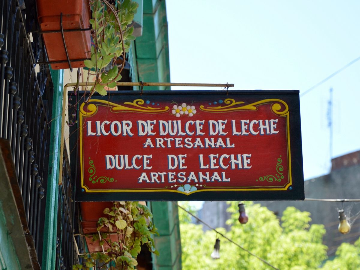 A wooden sign with a decorative border reads "Licor de Dulce de Leche Artesanal" and "Dulce de Leche Artesanal." It's hanging outside a building with potted plants.