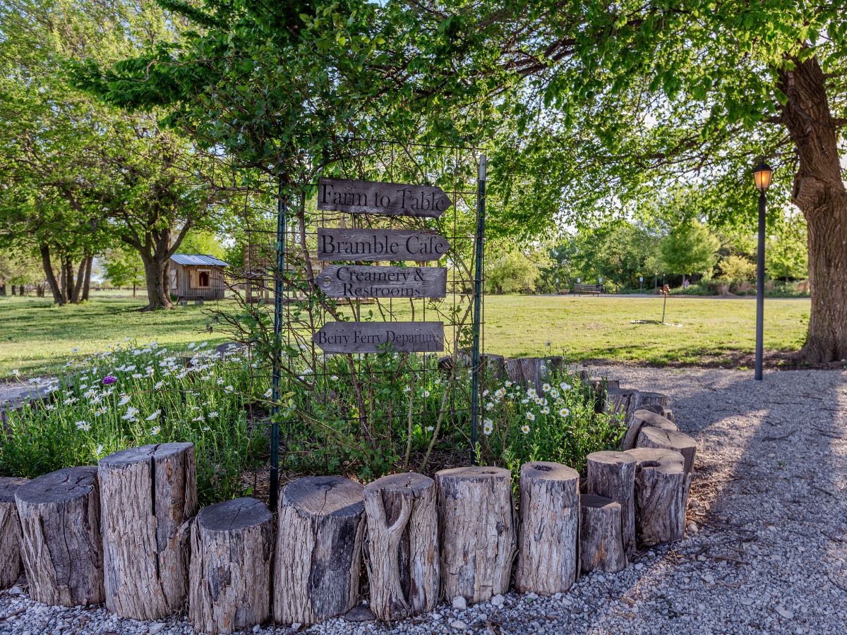 A wooden sign in a rustic garden reads "Farm to Table Bramble Cafe, Creamery, Restrooms, Berry Farm Pavilion." The area features wooden log borders, greenery, and a distant view of a building.