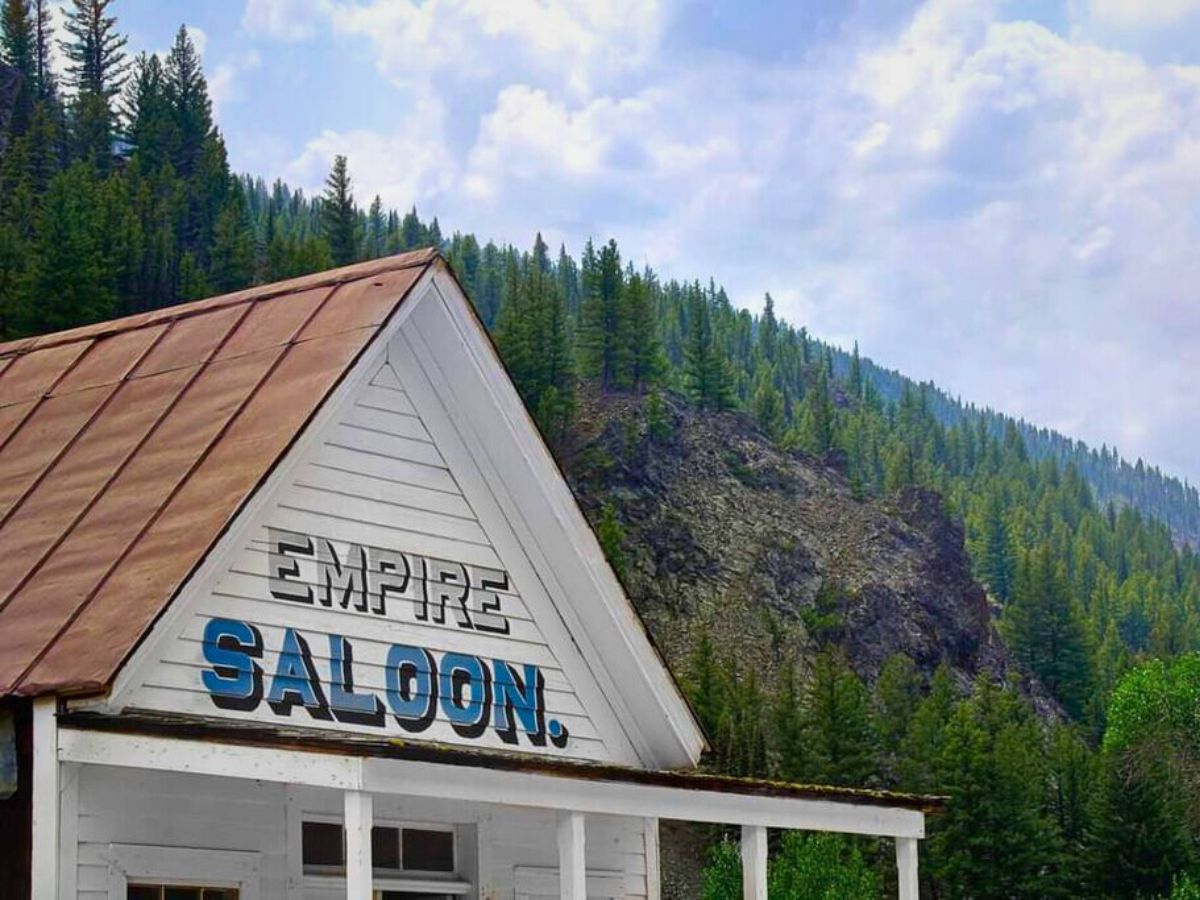 Empire Saloon building with a rusted metal roof, set against a backdrop of evergreen trees and rocky hills under a cloudy sky.