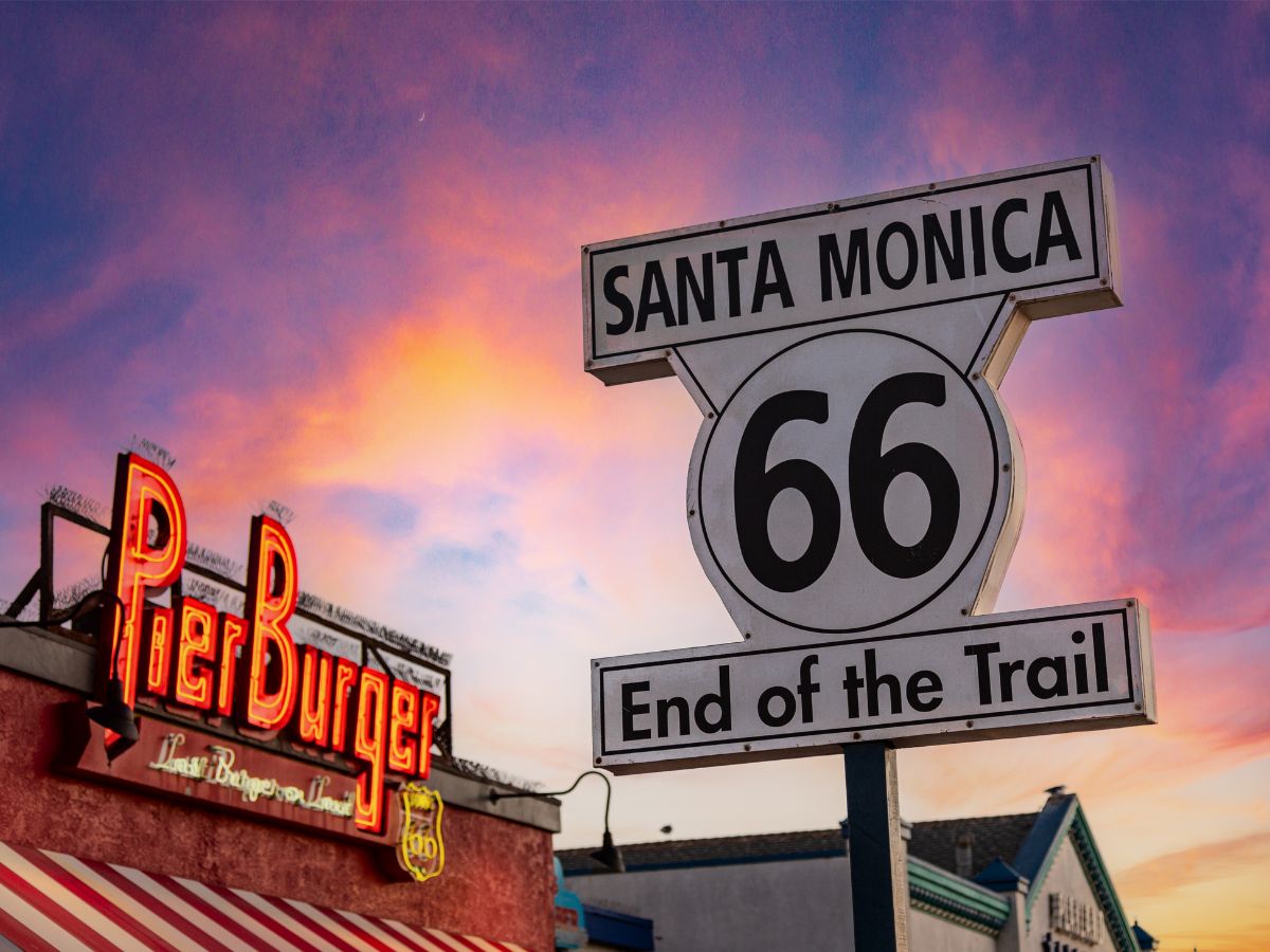 Route 66 "End of the Trail" sign next to a Pier Burger restaurant sign at sunset in Santa Monica.