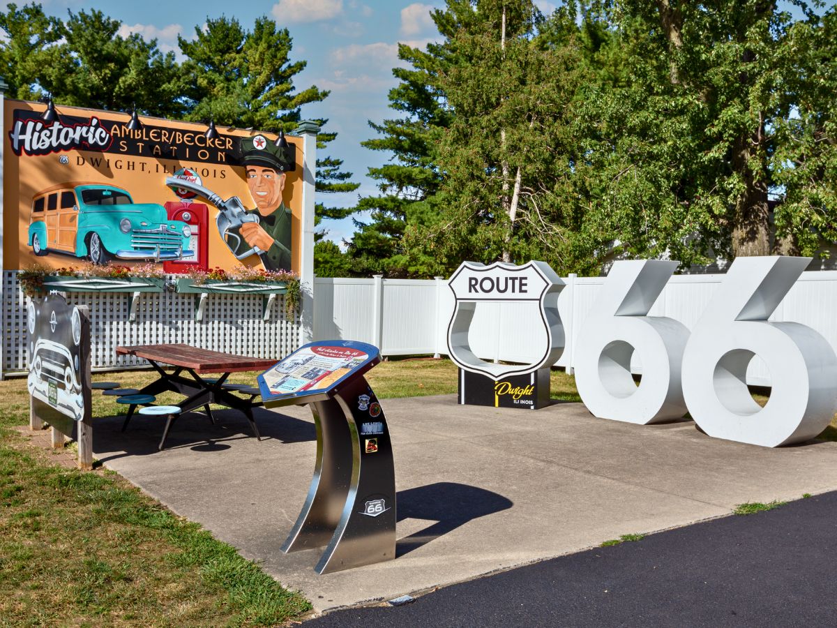 Route 66 sign display in front of a historical gas station mural, with a picnic table on a paved area and trees in the background.