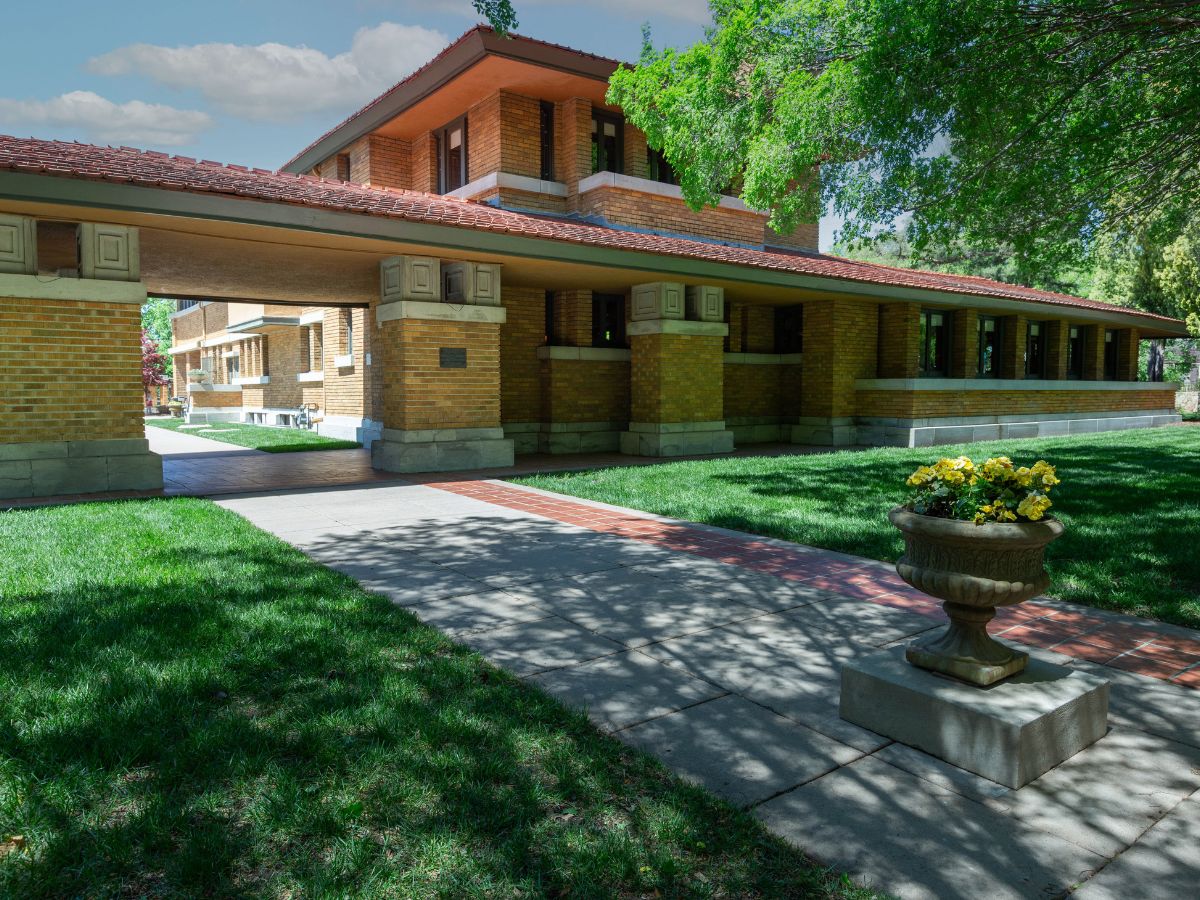 A two-story brick building with a red-tiled roof and large overhangs, surrounded by greenery, with a flower-filled planter on a concrete pathway in the foreground.