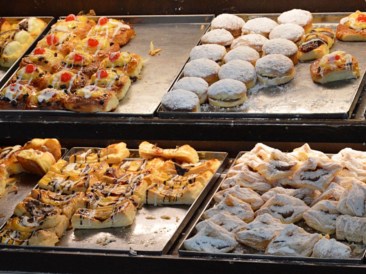 A bakery display showing trays of assorted pastries, including cherry-topped danishes, powdered sugar-covered donuts, and pastries drizzled with chocolate and icing.