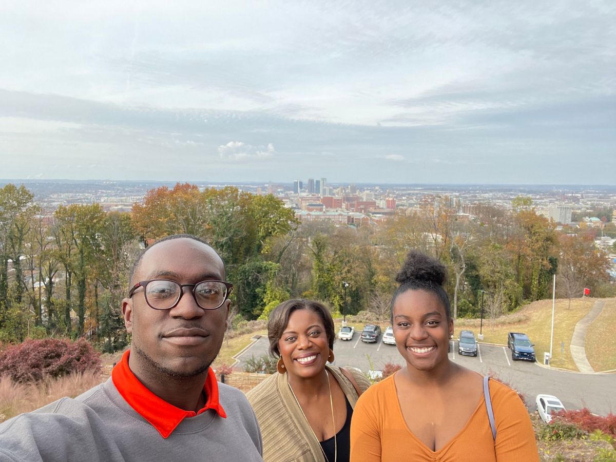 Three people smiling and standing together in front of a scenic view of a city landscape with trees and buildings in the background.