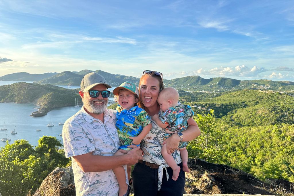 A man, woman, and two children, standing together atop a hill with a scenic view of a bay, boats, and rolling hills in the background.