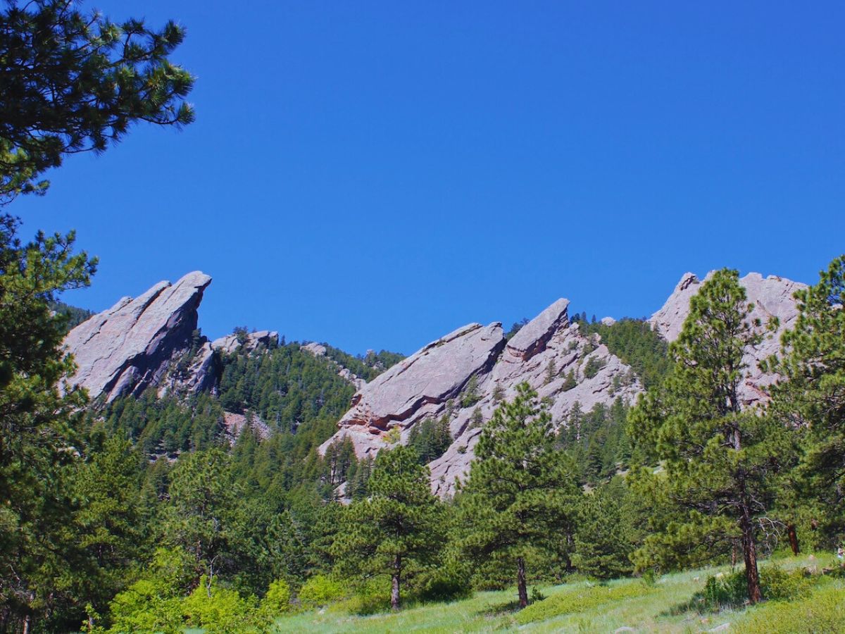 Rocky mountain formation with large, slanted flatirons surrounded by green trees under a clear blue sky.