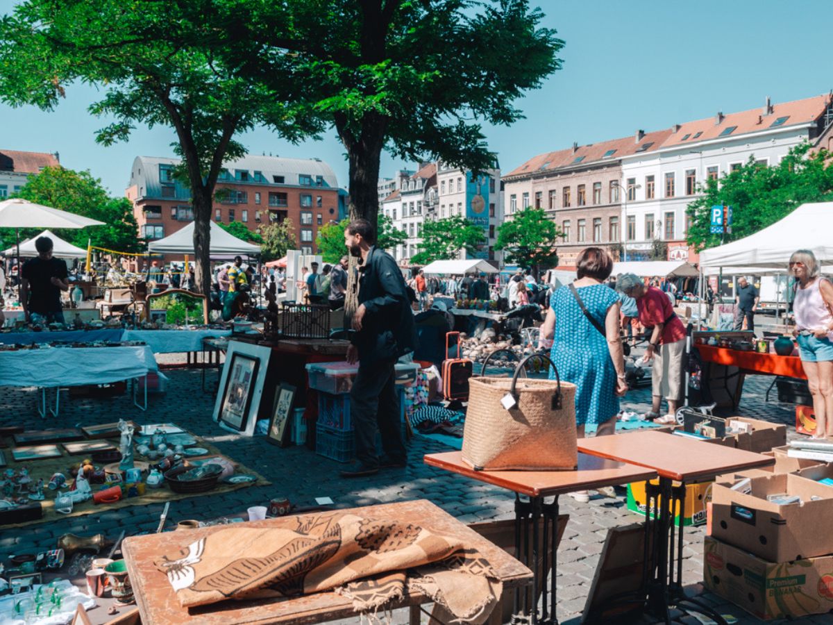 People browse various stalls at an outdoor market on a sunny day, with tables displaying a mix of items including pottery, framed pictures, and baskets.