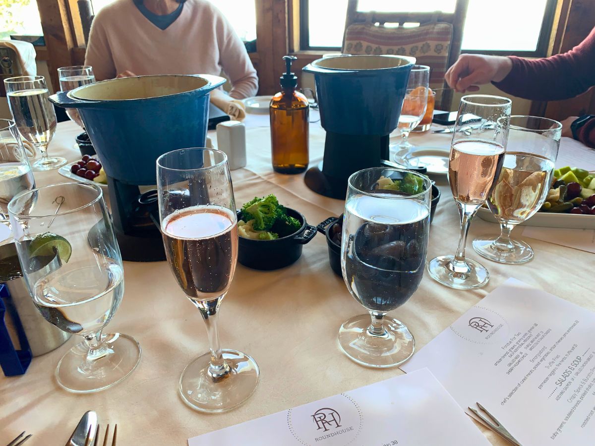 Table setting with fondue pots, glasses of water and sparkling wine, fruit bowls, and menus on a cream tablecloth. People are seated around the table.