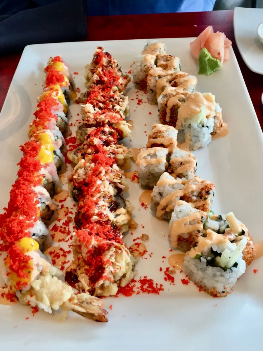 A white rectangular plate with three rows of sushi rolls topped with various toppings and sauces, including pieces of mango, red seasoning, and wasabi on the side.