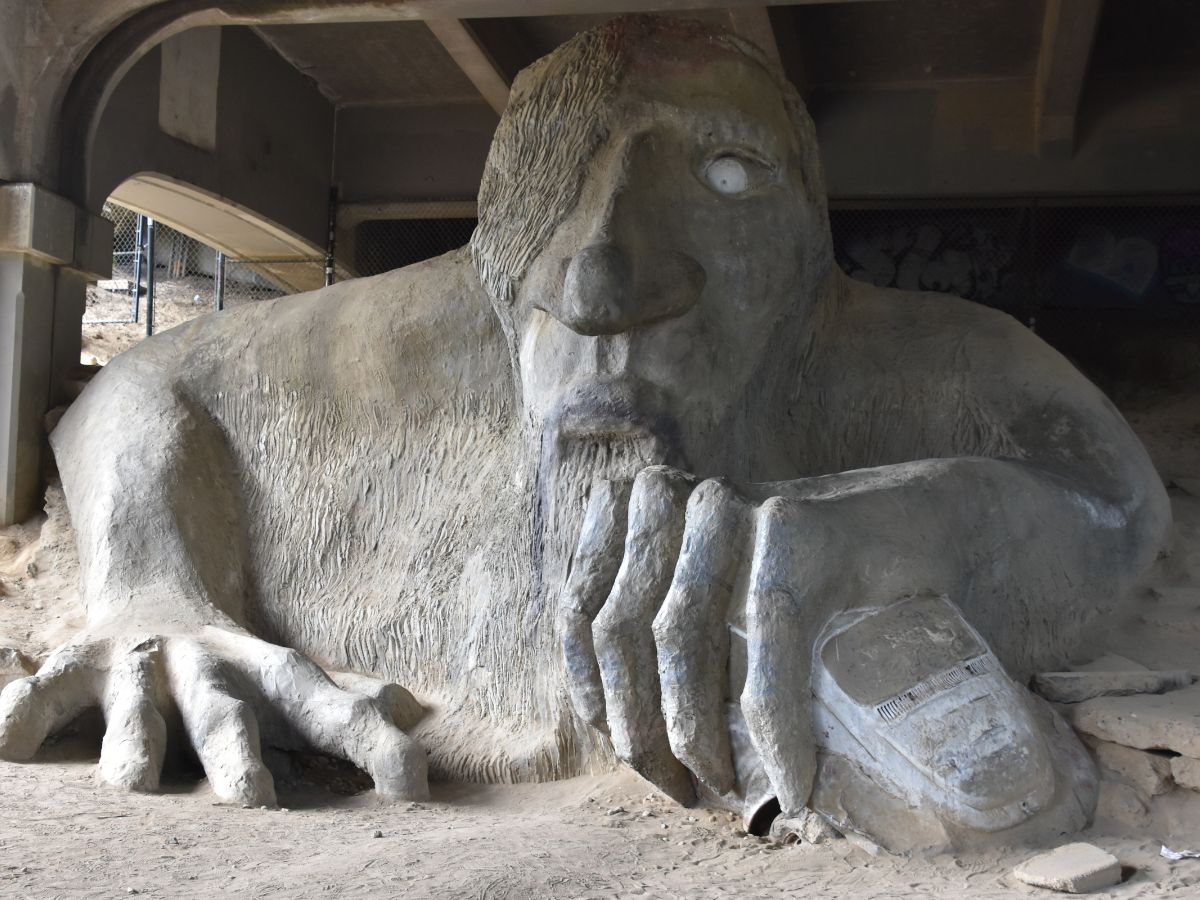 A large concrete sculpture of a troll grasping a car under a bridge. The troll has a rugged appearance and its single visible eye stares ahead.