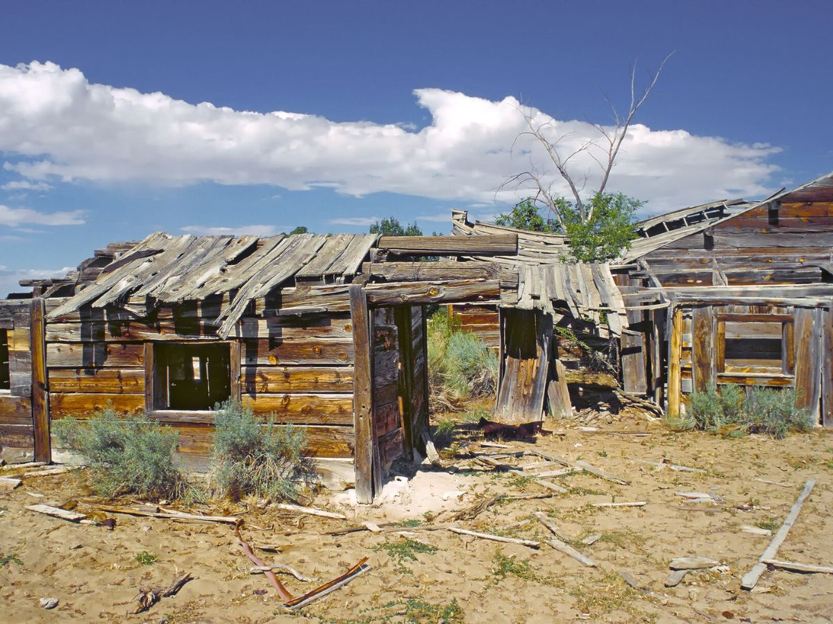 A weathered, abandoned wooden building with a collapsing roof, set in a sandy area under a partly cloudy sky. Sparse vegetation surrounds the structure.