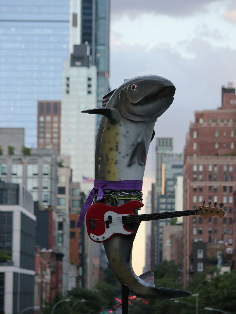 A sculpture of a leaping fish adorned with a purple scarf and holding a red electric bass guitar, set against a backdrop of tall city buildings.