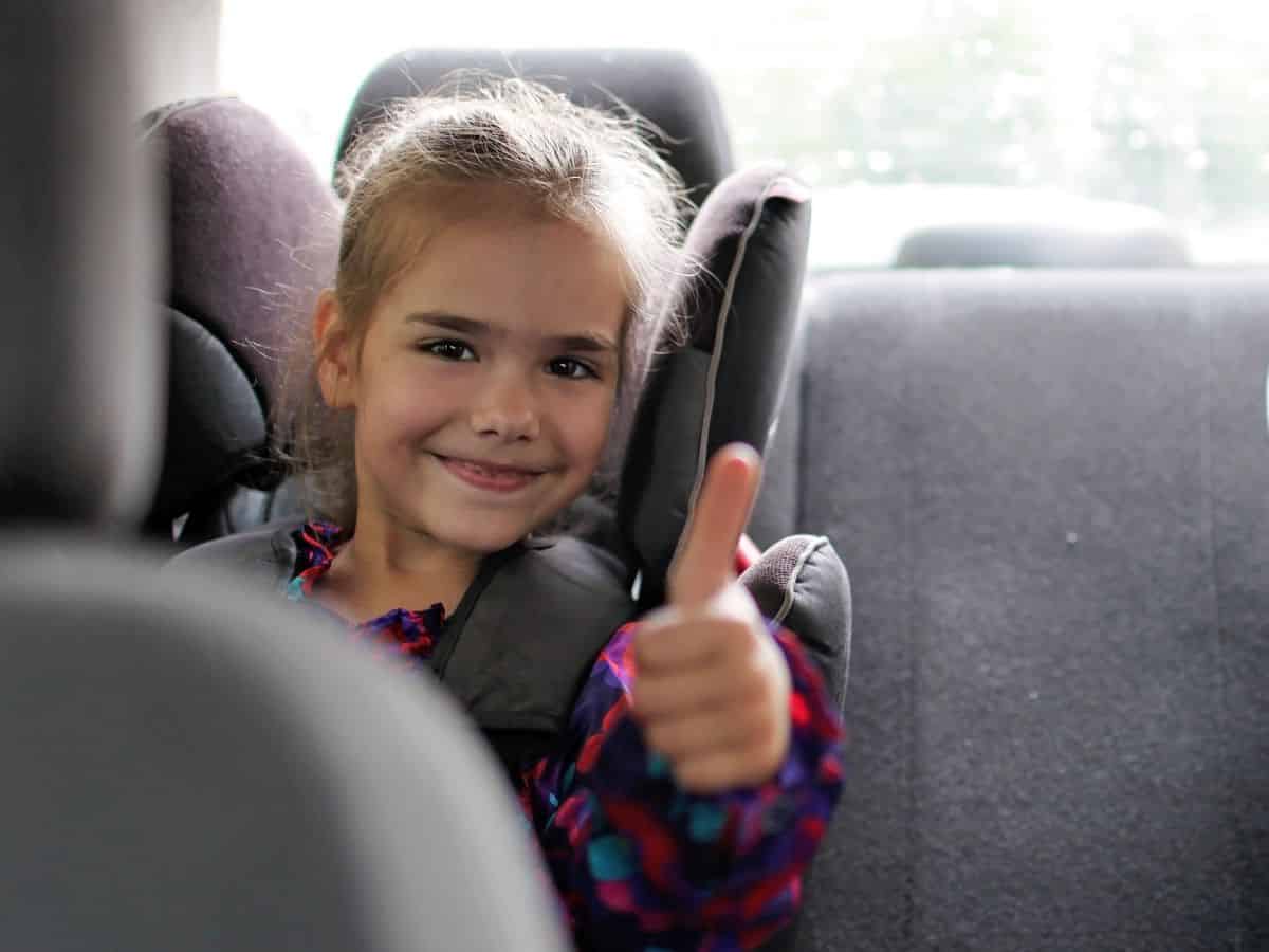 A little girl giving a thumbs up in a car seat enjoying family road trips with great audiobooks.