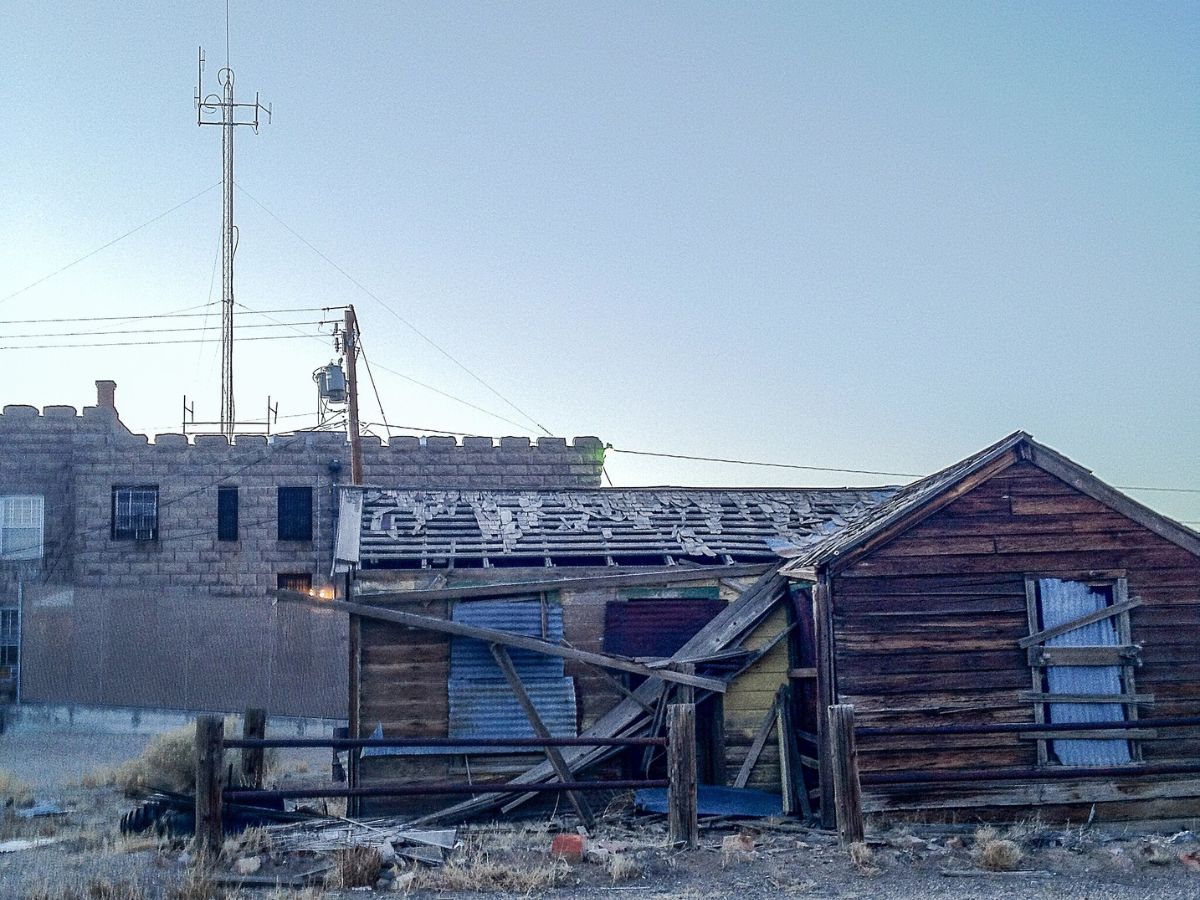 A dilapidated wooden building with a damaged roof stands in front of an older, castle-like brick structure with antennas on top under a clear sky.