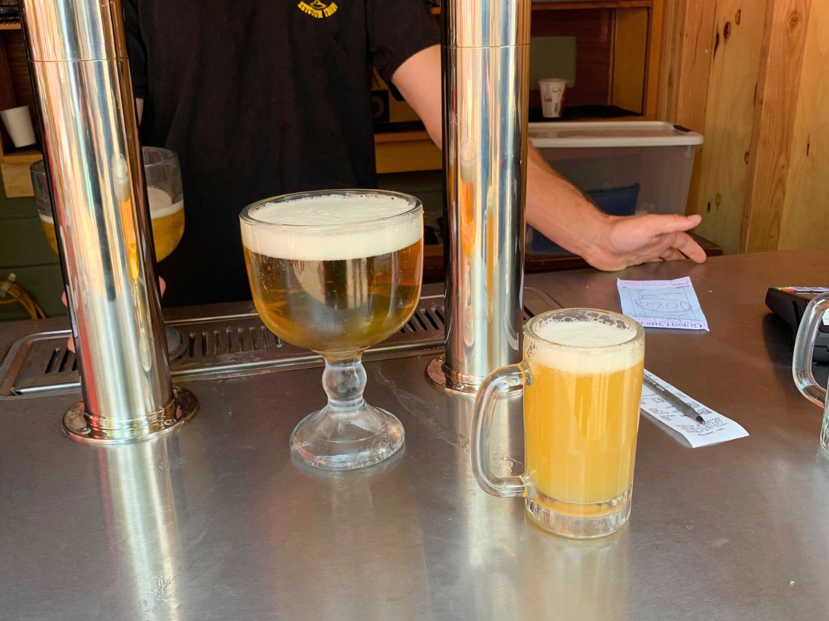 A bartender in a black shirt stands by beer taps with a large goblet and a mug of beer on the counter. A handwritten note and a payment terminal are on the right.