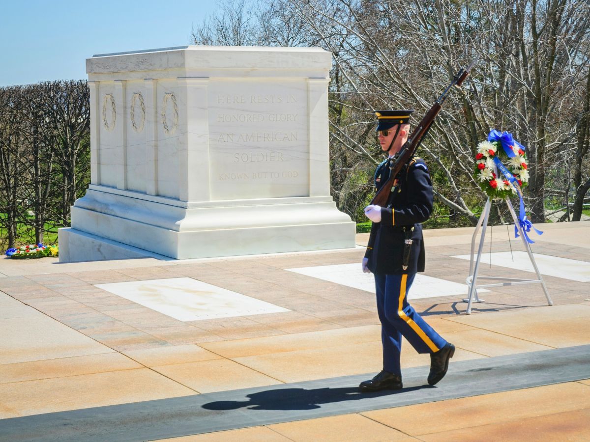 A guard walks beside the Tomb of the Unknown Soldier adorned with a wreath on a stand, surrounded by bare trees and a clear sky in the background.