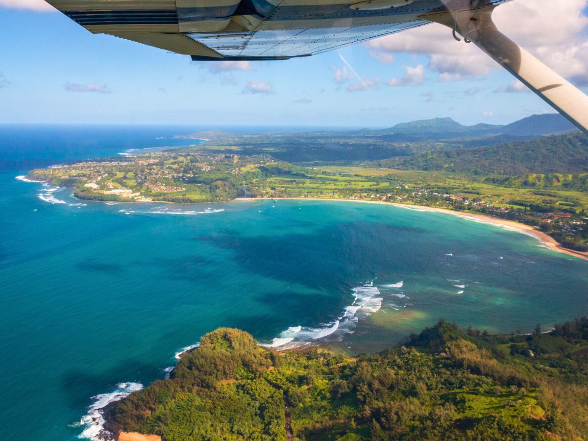 Aerial view of a coastline with a curved bay, lush greenery, and scattered clouds under a small plane's wing.