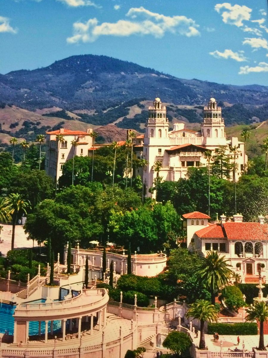 A grand estate with two towers surrounded by trees and rolling hills, under a blue sky with clouds.