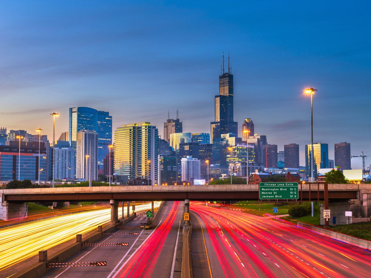 Evening view of a busy highway with light trails from vehicles, leading into a city with tall skyscrapers, including a prominent one with twin antennas.