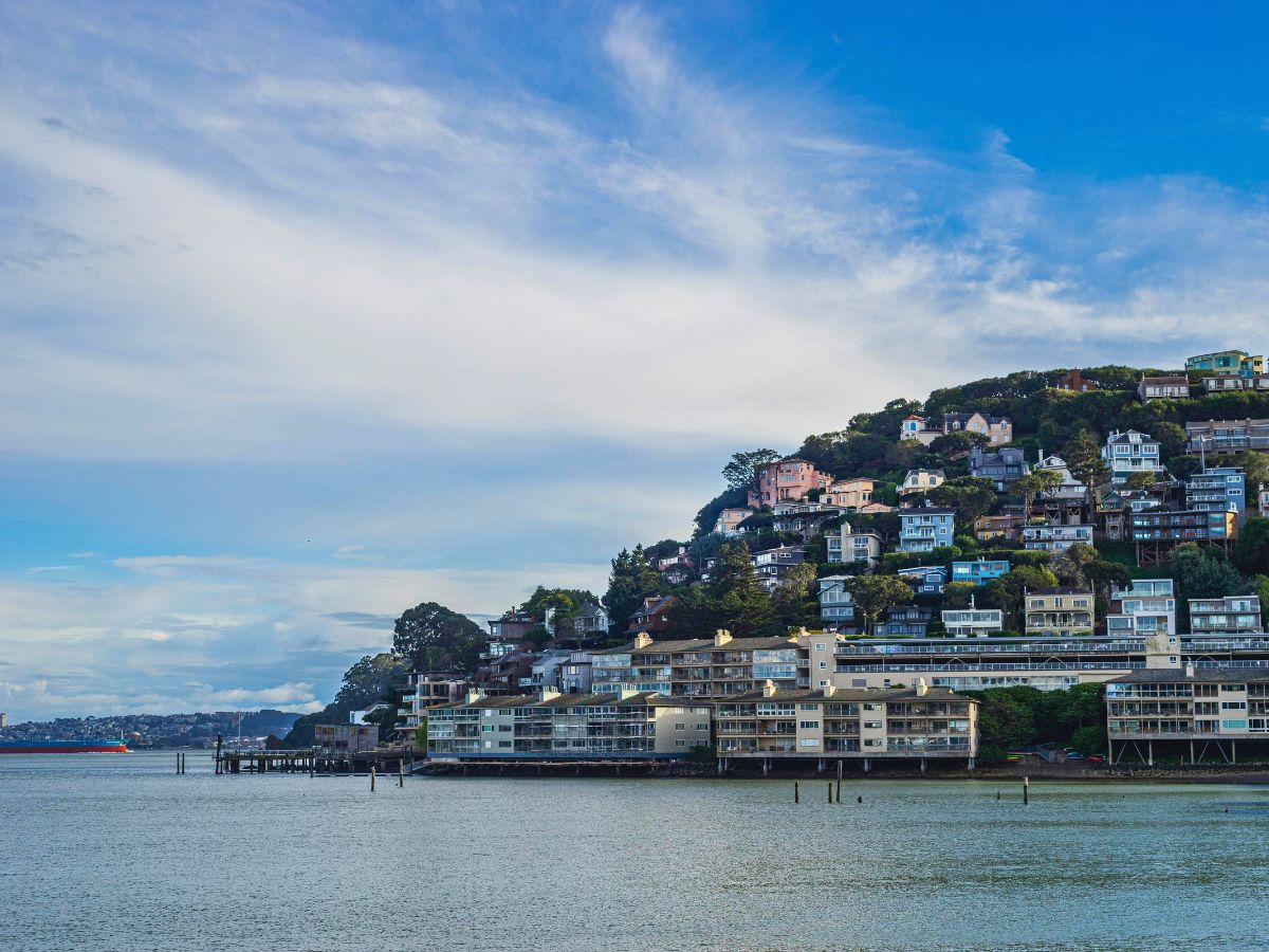 Hillside residential area with numerous houses overlooks a body of water, with a cloudy blue sky above.