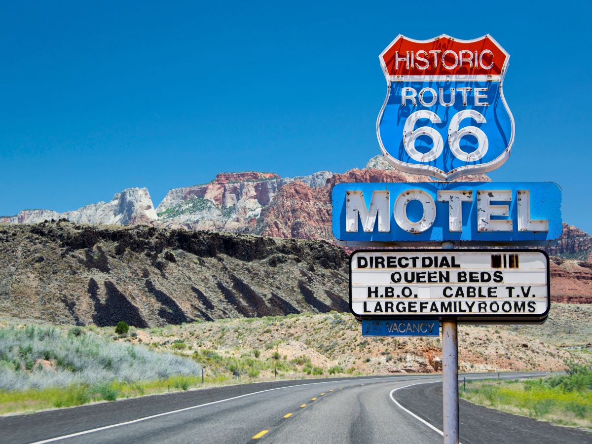 A vintage Route 66 motel sign in a desert landscape offering amenities like queen beds, HBO, and cable TV, with a mountainous backdrop.