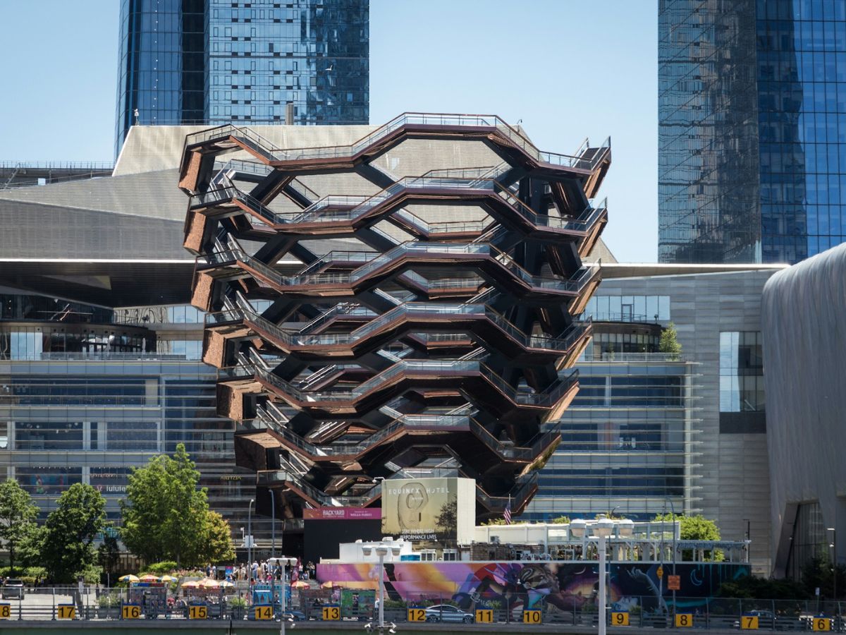 The image shows the Vessel, a honeycomb-like structure made of interconnected staircases, located in Hudson Yards, New York City. It stands between modern skyscrapers and other buildings.