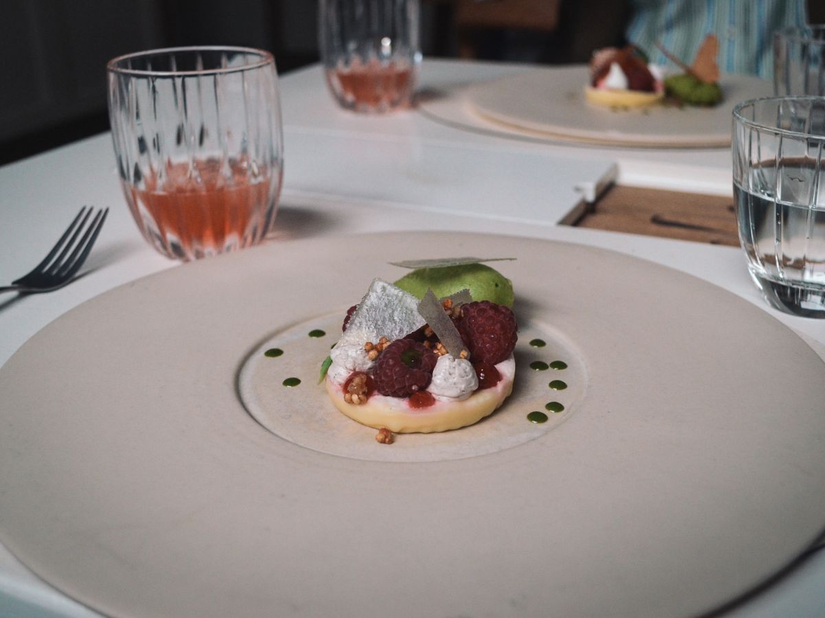 A desert plate featuring a cream tart topped with raspberries, edible garnish, and a small green scoop of sorbet, served on a white table with two glasses and a fork in the background.