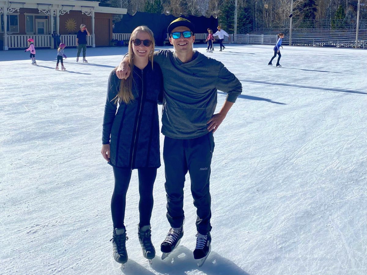 Two people stand on an ice rink, smiling and wearing ice skates. Other skaters and a building are visible in the background.