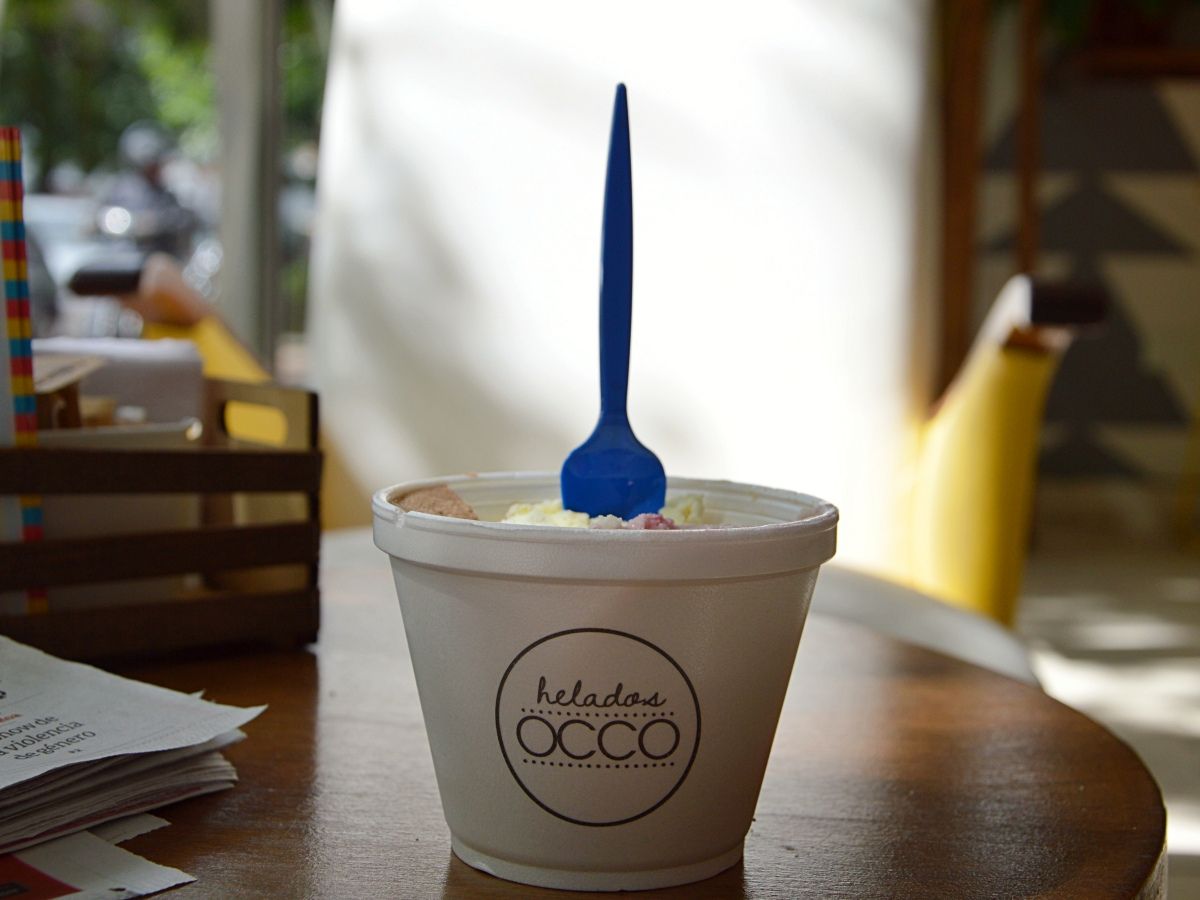 A cup of ice cream labeled "helados Occo" with a blue spoon inserted, placed on a wooden table in a sunlit room.